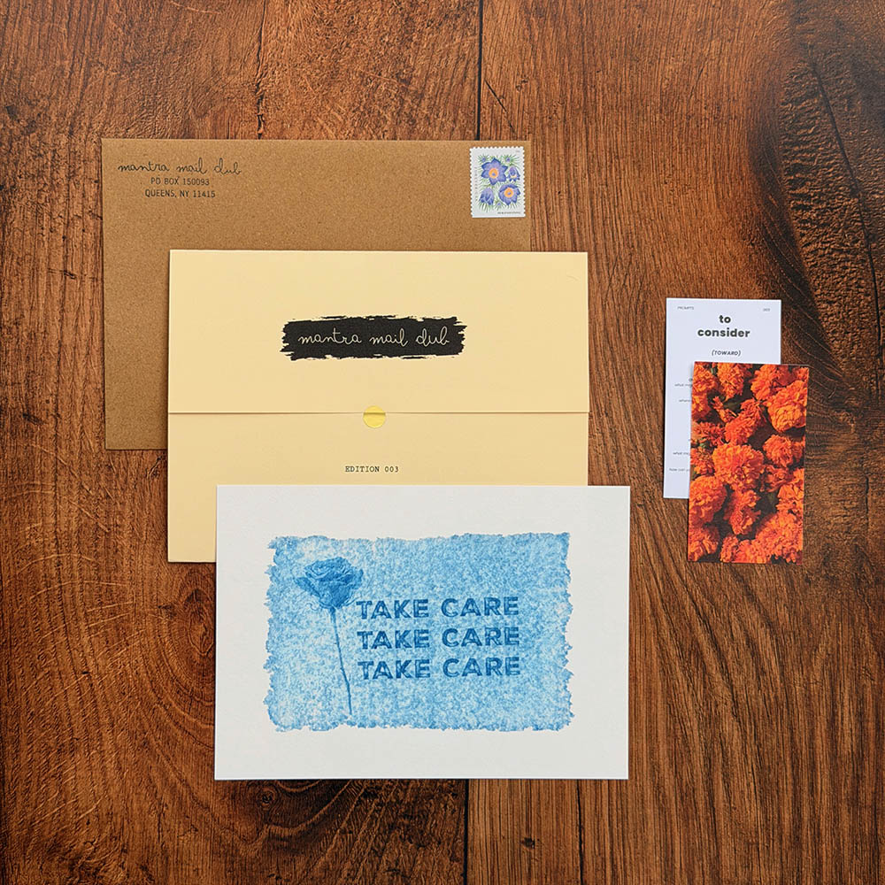 A flat-lay of a handmade greeting card set on a dark wood background, featuring a white card with blue watercolor text reading 'TAKE CARE', a yellow envelope with a black brush-stroke label, a brown kraft envelope, and a small orange floral card.